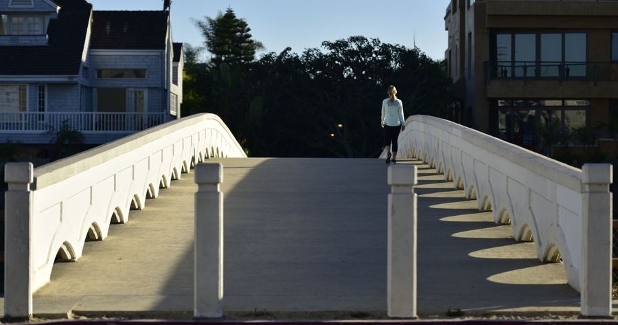 A white arched footbridge in low sun, with one person walking across it.