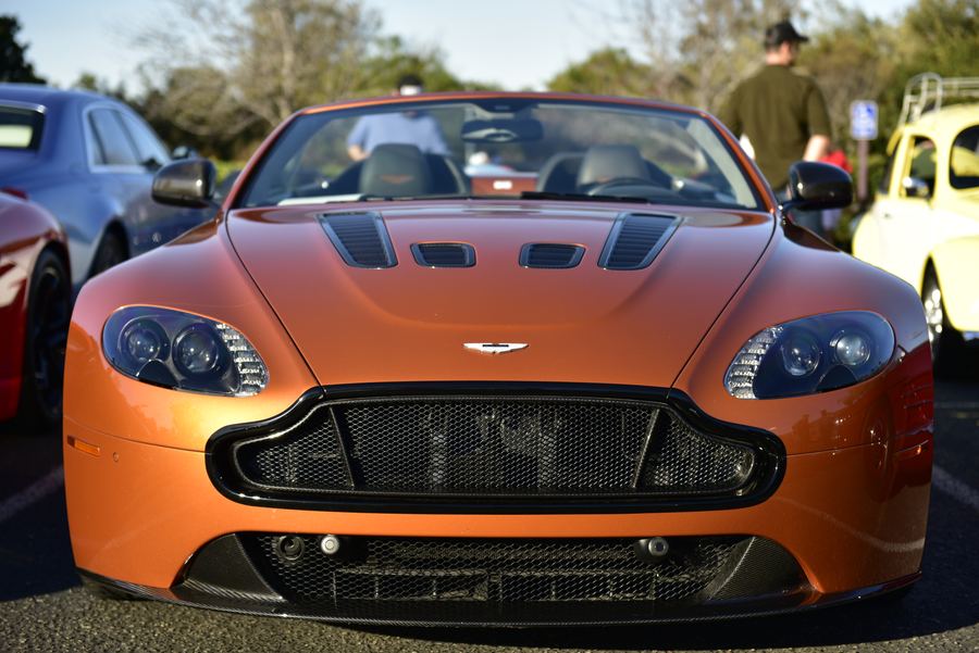 The front of an orange convertible sports car in a parking lot, with other cars and people around it.