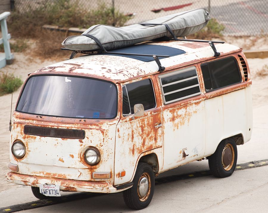 A rusted white vintage van with a roof rack, parked near a beach.