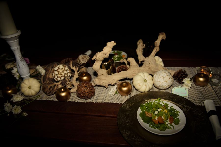 A dinner table at night with a driftwood and mini pumpkin centerpiece, gold ornaments, and a plated salad in the foreground.