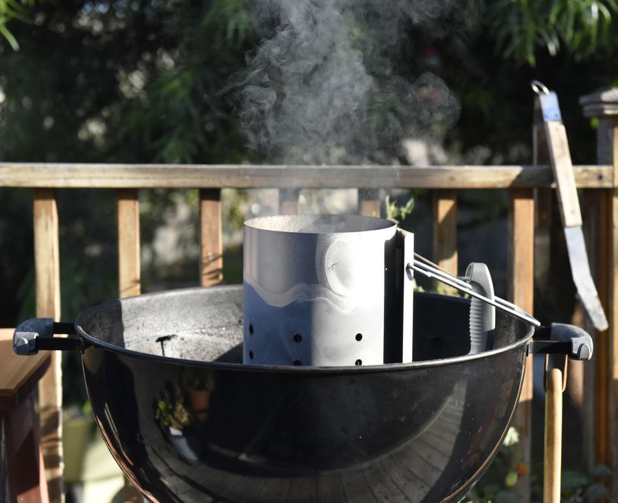 On a wooden deck, a chimney starter on a black kettle grill releases smoke.