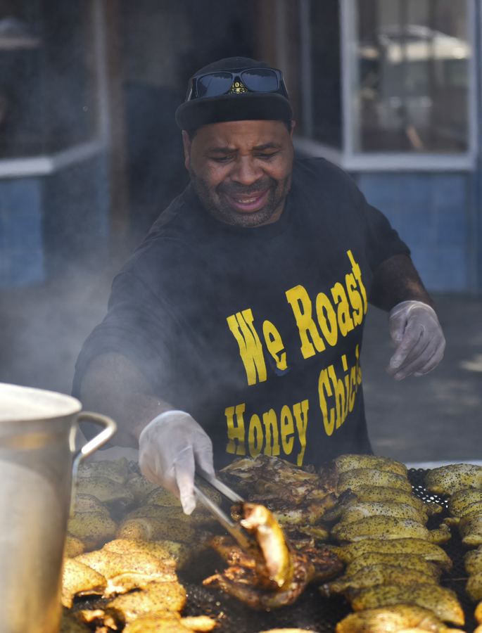 A cook in gloves turns chicken on a smoking outdoor grill. The cook's black shirt says We Roast.