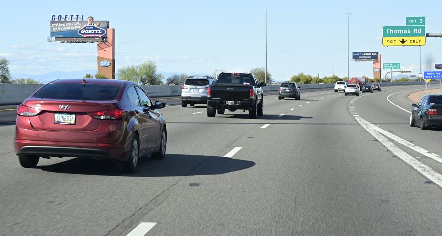 Freeway traffic heading toward billboards and green exit signs under a blue sky.