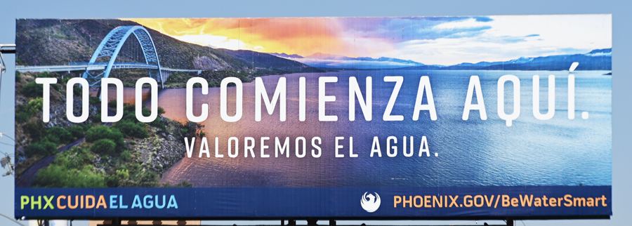 A Spanish-language billboard for the Phoenix water campaign, with a lake and bridge at sunset in the artwork. It reads Todo comienza aquí, valoremos el agua.