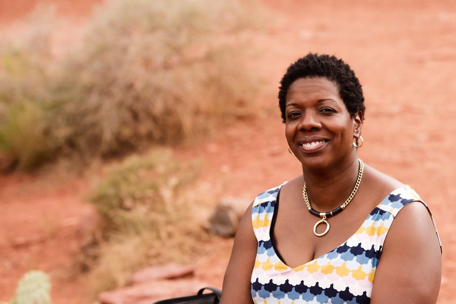 A smiling woman in a patterned sleeveless top, with short natural hair, outdoors against red desert soil and brush.