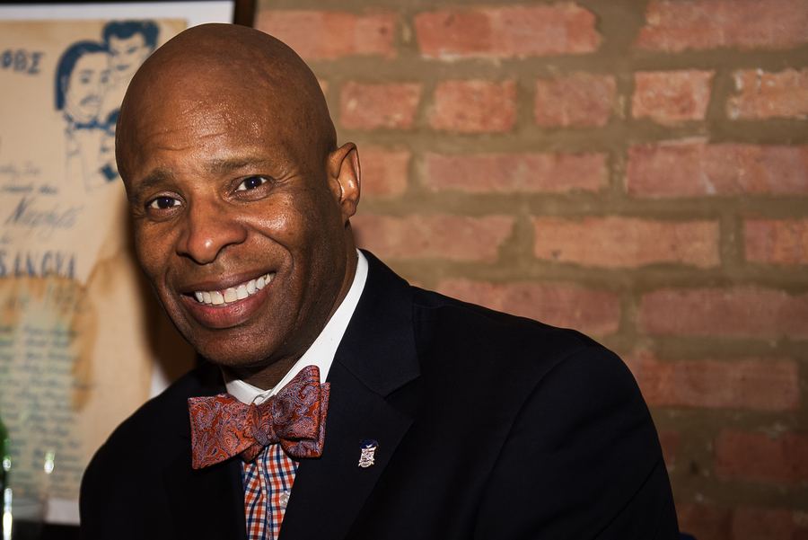 A smiling bald man in a dark suit and patterned bow tie in front of a brick wall.