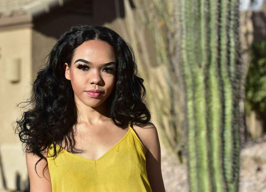 A young woman in a yellow top, with long dark curls, photographed outdoors beside a saguaro cactus.