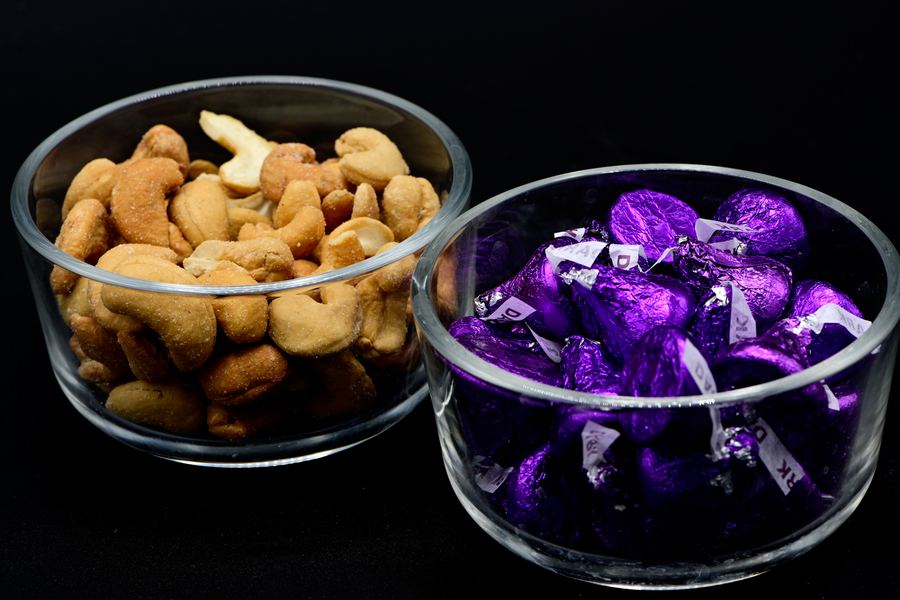 Two glass bowls on a black background. One holds cashews. The other holds chocolates in purple foil.