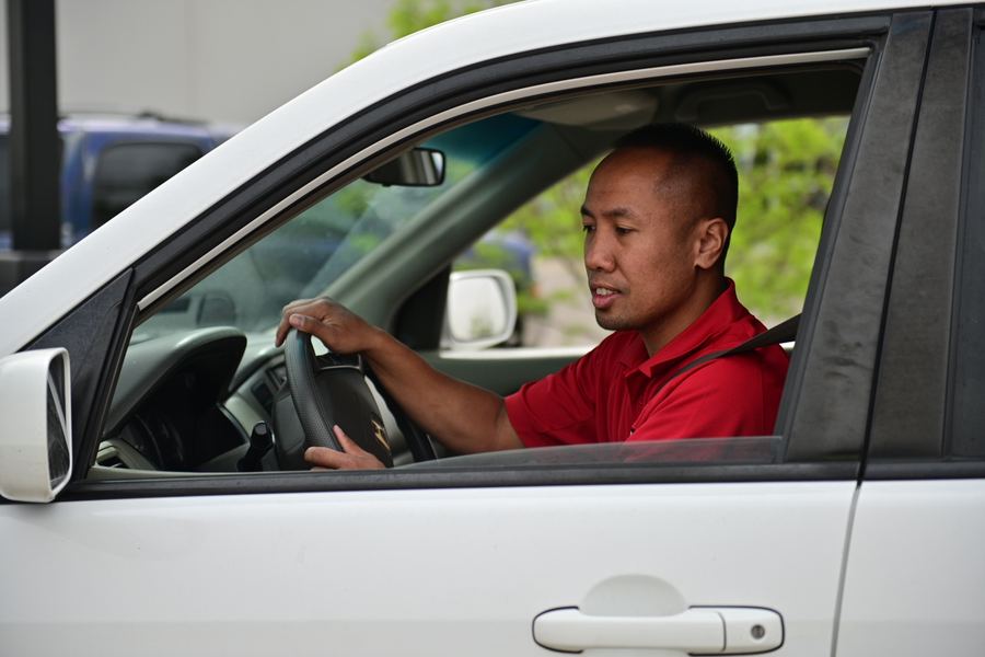 Seen through the open window, a man in a red polo sits in the driver's seat of a white SUV with both hands on the wheel.