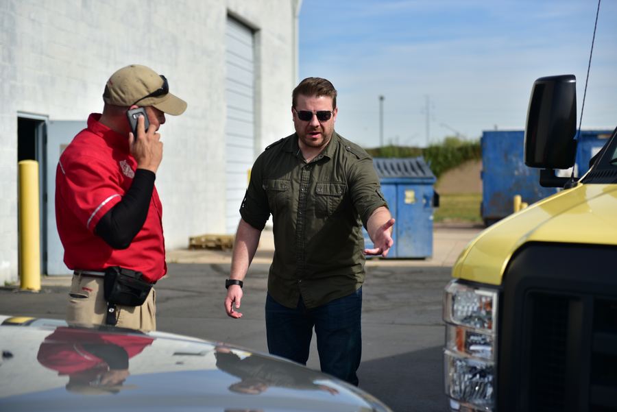 Two men talk beside a yellow delivery truck in a parking lot. One wears a red shirt and cap and holds a phone to his ear.