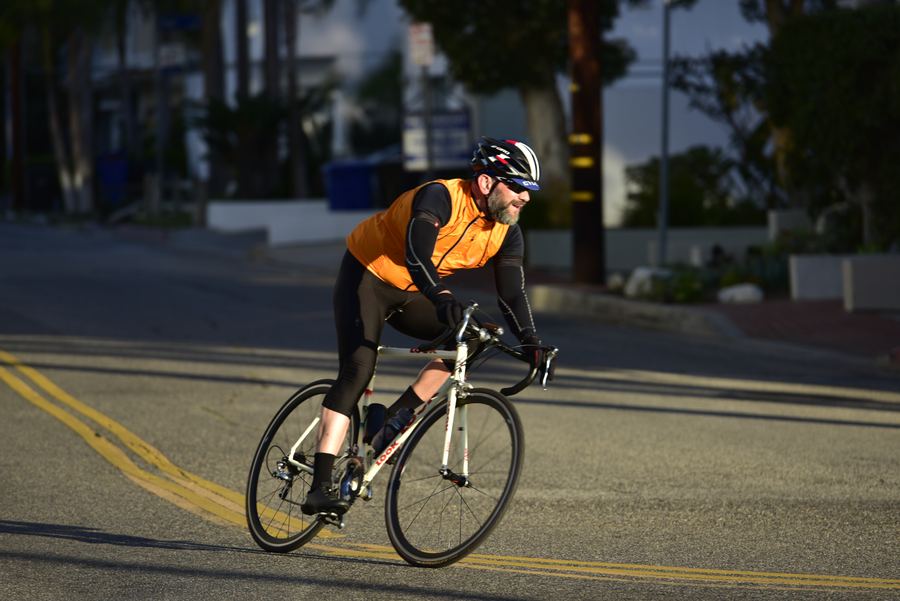 A cyclist in an orange vest and helmet leans through a turn on a street in low sun.