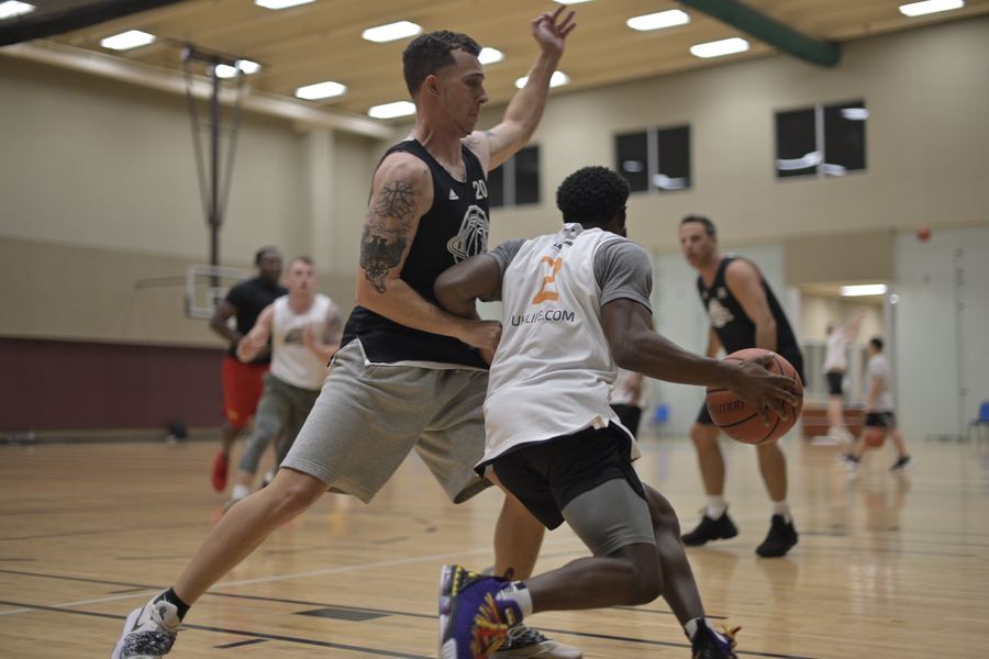A player in a white number 2 jersey drives with the ball as a defender in a black number 20 jersey leans into him. Other players run up the court behind them.