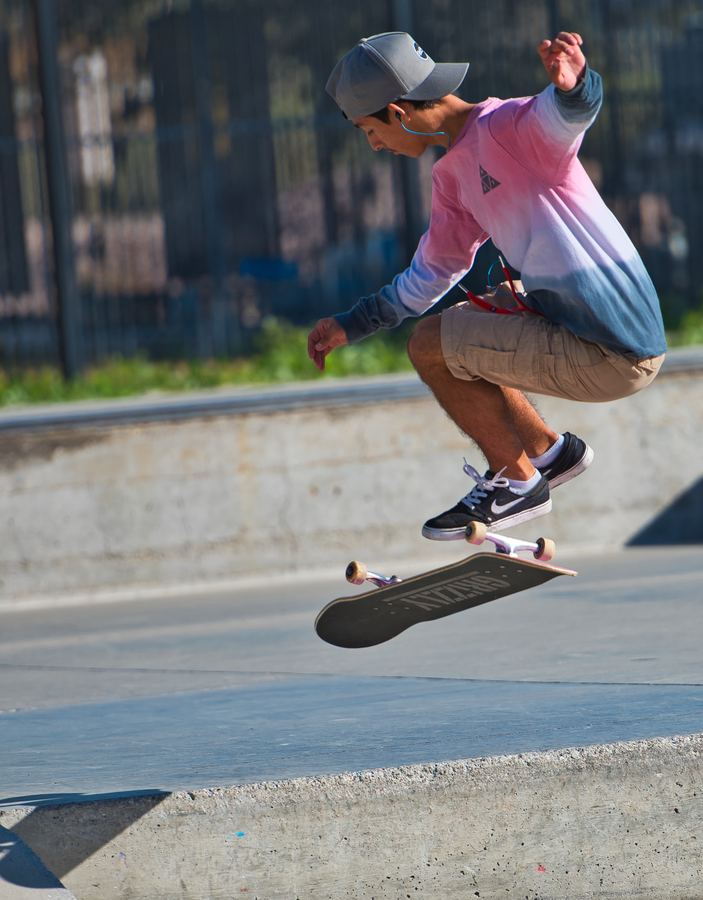 A skateboarder in a cap and a pink and blue sweatshirt jumps, with the board in the air beneath him.