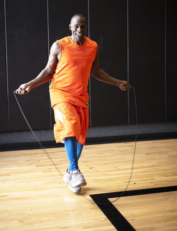 A man in an orange tank top and shorts jumps rope on a hardwood gym floor.