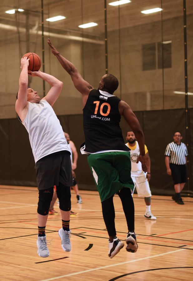 A player in a white jersey rises for a jump shot as a defender in a black number 10 jersey leaps with one arm up to block it. A referee watches from the back of the gym.