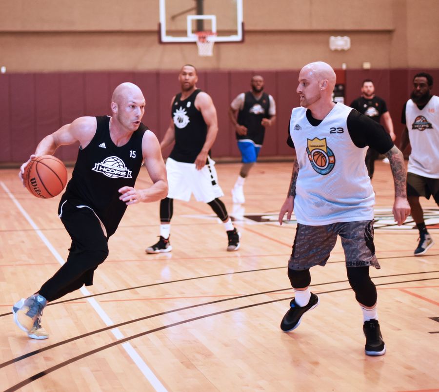 A player in a black number 15 jersey dribbles toward the lane. A defender in a white number 23 jersey sets his feet in front of him.