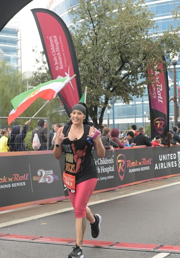A runner in pink leggings and a black tank top makes a hand sign as she crosses the finish line. Crowds and banners fill the background.