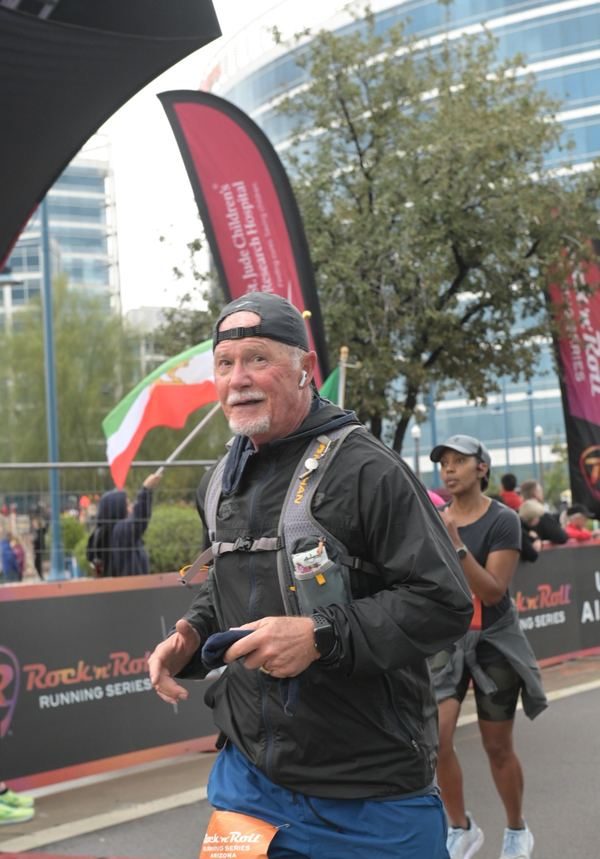 An older runner in a black jacket and backward cap glances up near the finish line as another runner follows.