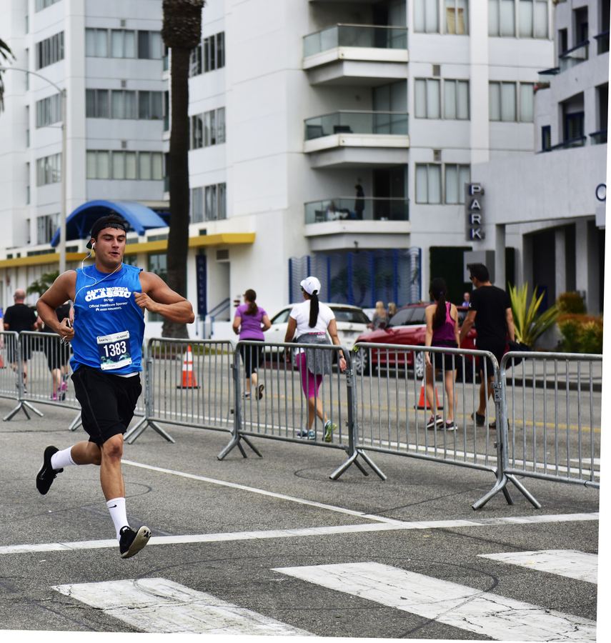A runner in a blue shirt and race bib crosses a street, with barricades and other runners behind him.