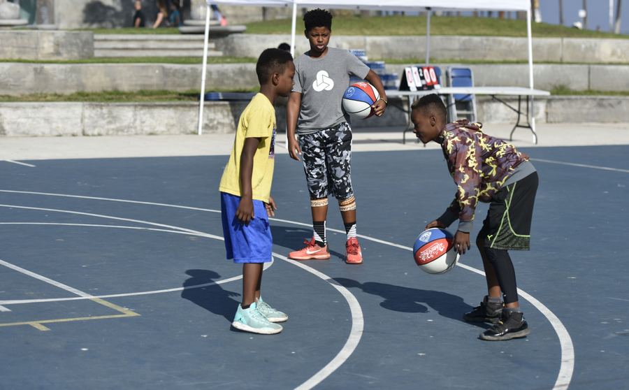 Three young people play basketball on an outdoor court. One dribbles a red, white and blue ball.