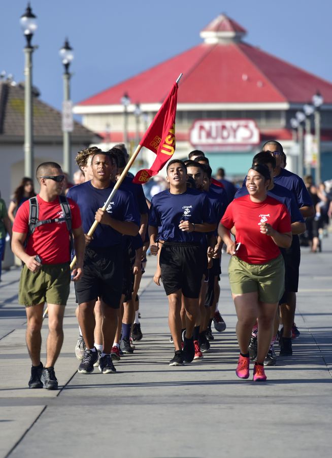 A group in red and navy shirts jogs along a pier behind a person carrying a red flag.