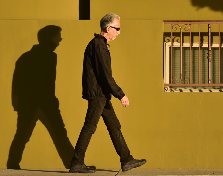 A man in dark clothes and sunglasses walks along a yellow wall with his shadow beside him.